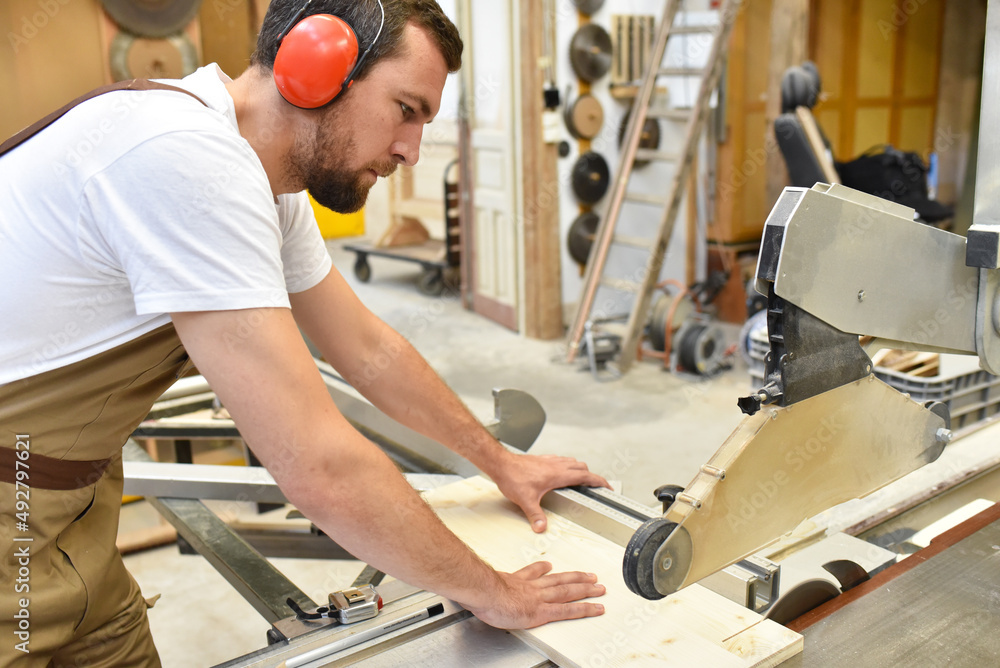 Wall mural young carpenter in working clothes works in the joinery on a sanding machine - working clothes with ear protection