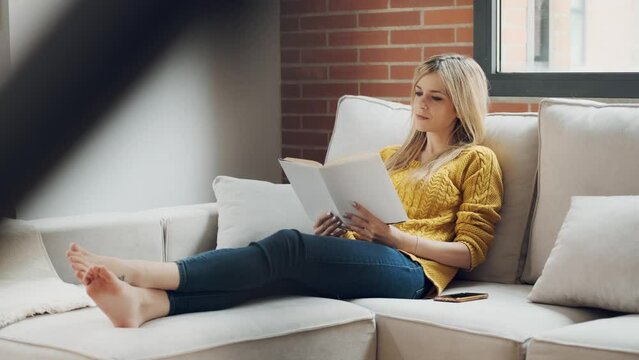 Video of pretty young woman reading a book while sitting on sofa at home.