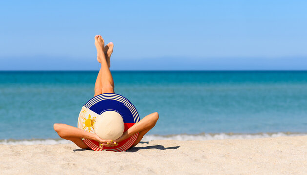 A slender tanned girl on the beach in a straw hat in the colors of the flag of the Philippines. The concept of a perfect vacation in a resort in the Philippines. Focus on the hat.
