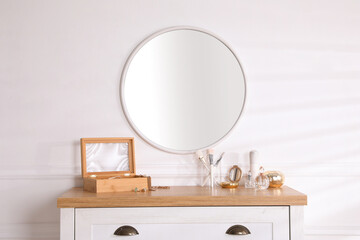 Trendy round mirror and chest of drawers with accessories near white wall indoors.