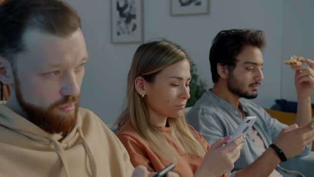 Diverse group of young people using smartphones eating pizza and watching TV at night in dark living room. Modern lifestyle and youth culture concept.