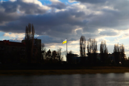 Yellow-blue flag of Ukraine flying against overcast sky. Symbol of Ukrainian freedom, independence a sign of hope. City view with trees, silhouettes of houses with tilt-shift effect. War Ukraine 2022.