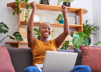 A young woman celebrates a big win by raising her arms on the couch, with laptop - obrazy, fototapety, plakaty
