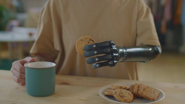 Cropped shot of unrecognizable girl with prosthetic arm holding tea mug and cookie while sitting at kitchen table