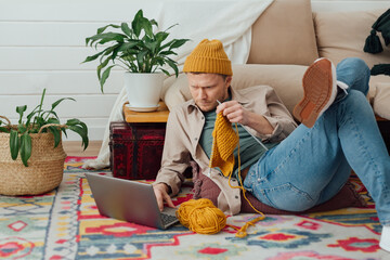Young serious man looking at laptop. Man learning new hobby, knitting on needles. Knitting project in progress. - 492821038
