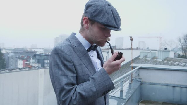 In this video is a nice suited man who starts to smoke his pipe on the balkony, while small snowflakes are coming down in the backround. He&acute;s wearing a grey beret and a handsome mustache.