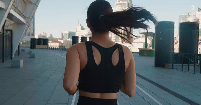 Rear back view of athletic young woman jogging outdoors in slow motion. Fitness girl doing cardio workout, running in the morning on city streets