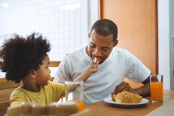 Fototapeta na wymiar Black African American little boy feeding Croissant or bread father at dining table in the kitchen together.