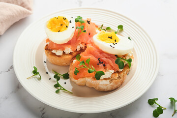 Sandwich with delicious toppings, smoked salmon, eggs, herbs and microgreens radish, black sesame seeds over white plate on white marble table background. Healthy open sandwich superfood. Top view.