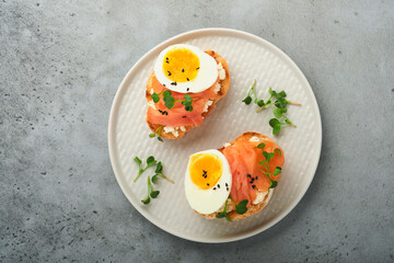 Sandwich with delicious toppings smoked salmon, eggs, herbs and microgreens radish, black sesame seeds over white plate on gray concrete table background. Healthy open sandwich superfood. Top view.