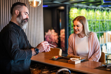 Sushi master is teaching young woman how to use chopsticks at sushi restaurant.