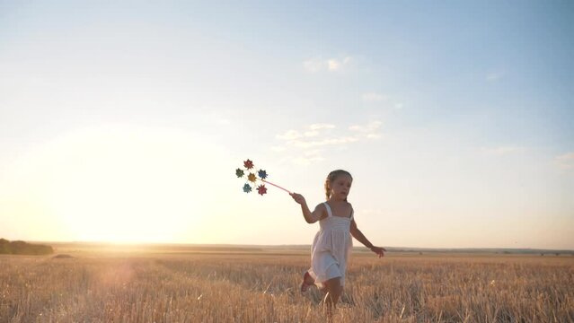 Happy girl runs across field at sunset with windmill in her hands. Silhouette of joyful girl holding wind toy. child enjoys fresh air. fun game in wind in field. Happy child runs at sunset.Wind toy
