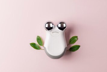 microcurrent facial toning device on pink background with green leaves. Modern beauty skincare...