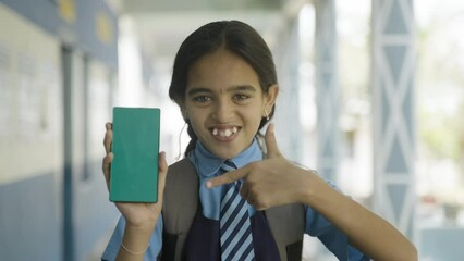 Smiling school girl kid in uniform showing green screen mobile phone by pointing finer while looking camera at corridor - concept of education advertisement and learning application promotion