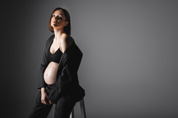 Trendy pregnant woman in jacket posing near chair on dark grey background