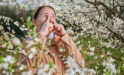 Woman allergic suffering from seasonal allergy at spring, posing in blossoming garden at springtime. Young woman sneezing among blooming trees. Spring allergy concept - obrazy, fototapety, plakaty