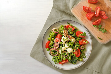 Healthy vegetarian salad from green vegetables with feta cheese, braised onions and tomatoes on a plate, napkin and cutting board on a white table, copy space, top view from above