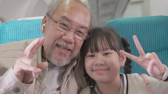 Happy Asian grandfather and granddaughter showing peace sign finger and mini heart looking at camera at seat in airplane cabin. family trip travel vacation