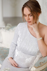 beautiful pregnant woman in evening dress sits on a sofa in a white room. The concept of motherhood and the birth of children. Glamor and beautiful life