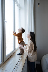 A mother and a small child stand at a large white window and look out into the street. Safety for small children in the house. - obrazy, fototapety, plakaty