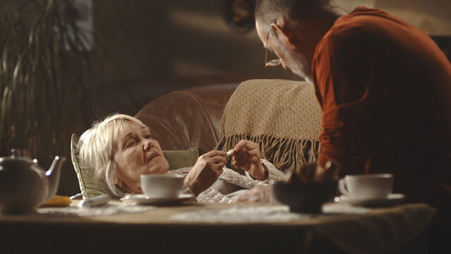 Elderly husband checking digital thermometer and speaking to sick wife in cozy living room at home - Powered by Adobe