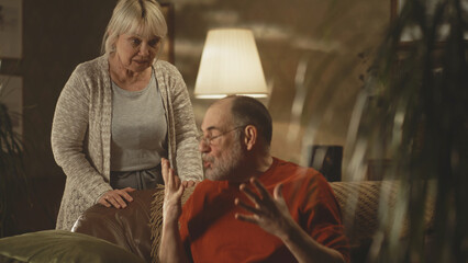 Rack focus of irritated senior husband sitting on sofa and shouting at angry wife during quarrel in living room at home