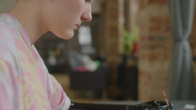 Selective focus shot of young Caucasian girl with prosthetic arm sitting at table and painting picture on paper with brush at home