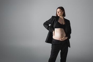 Stylish pregnant woman in top and jacket touching belly isolated on grey