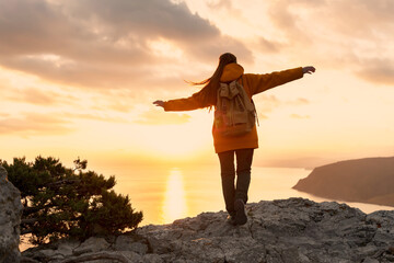Fototapeta premium Female hiker walks with backpack on top of mountain. Girl stands with raised arms at sunset and enjoys freedom