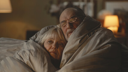 Upset elderly man discussing gas crisis and war with sad wife while wrapping in warm blanket on couch in dim cold living room at home - obrazy, fototapety, plakaty