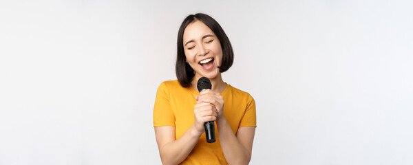 Happy asian girl singing and having fun, holding microphone at karaoke, standing in yellow tshirt...