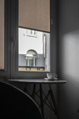 A lone white cup and saucer on a white folding table by the window in a hostel room opposite the illuminated wall and windows of the house next door. The concept of loneliness and minimalism