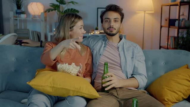 Joyful couple watching sitcom laughing eating popcorn and drinking beer in apartment at night. Relationship and leisure activity concept.