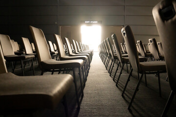 Low angle shot, front view of a distant opened door with a sign above written in both English and Thai means EXIT, with bright light shone into the hall through the aisle between rows of empty chairs. - obrazy, fototapety, plakaty