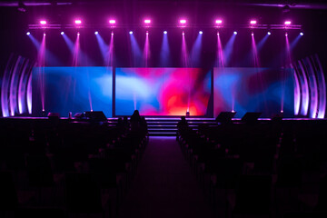 Front view of the stage event with a big LED screens and purple and pink lights are shining down on the stage floor while testing the light system with many empty chairs arranged for audience in hall. - obrazy, fototapety, plakaty