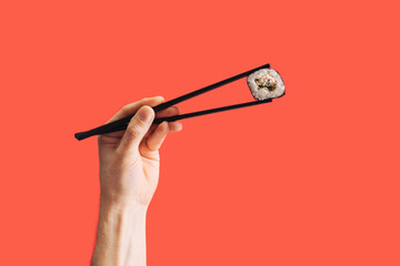 Sushi vegetarian eating minimal concept. Man hand holding black chopstick with one piece of squid and rice rolled in nori dried edible seaweed. Bright red background