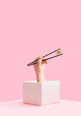 Takeaway sushi minimal concept. Man hand holding black chopstick pop up from box with one piece rice salmon and cucumber roll in nori dried edible seaweed. Baby pink background