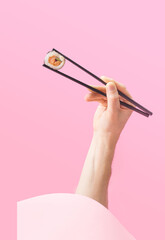 Sushi takeaway minimal concept. Man hand holding black chopstick pop up from round box with one piece rice salmon and cucumber roll in nori dried edible seaweed. Awesome pink background