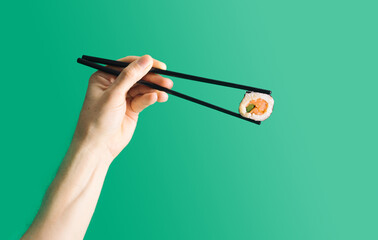 Minimal flat lay sushi concept. Man hand holding black chopsticks with one piece, salmon fish, rice, cucumber and nori seaweed. Pastel green background