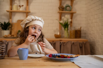 a cute six-year-old girl in a chef's hat and apron is sitting at a wooden table eating pancakes