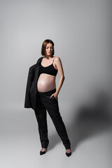 Full length of stylish pregnant model holding jacket on grey background