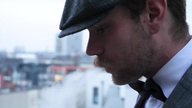 There is a well dressed man with a grey suit and a grey beret, a 3 day beard and a lovely mustache. He is smoking a cigarette on a grey and cold wintersday. Obviously he enjoy&acute;s his smoking break.