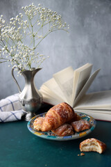 Still life with freshly baked croissants on a plate, an open book, white gypsophila flowers in vintage silver vase. Weekend breakfast concept. 