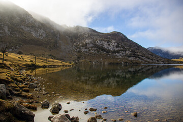 Fototapeta premium Paisaje montañoso con neblina, adornado en la orilla de un lago pedregoso. Cielo azul y reflejos del lago .Lagos de Covadonga 