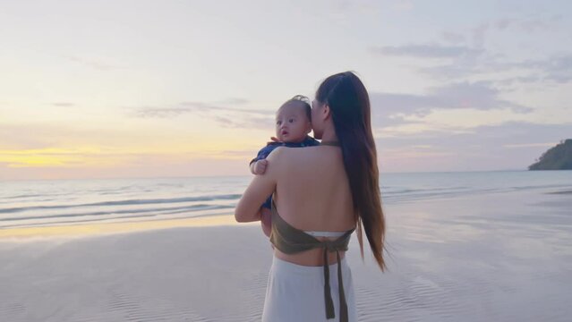 Happy mother with baby boy walks by ocean on the beach in summer. Both are having fun time and cheerful together. Motherhood love and care for her child. An amazing view retreats them a lot.