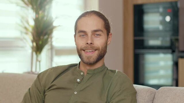Face of attractive hipster guy wearing casual colothes ponytail smiling to camera indoors. Male portrait. Attractive man staying home. Apartment.