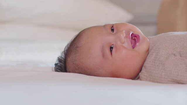 An Asian newborn baby is laying down on a soft white sheet mattress. Starring at his parents and feels sleepy. Adorable chubby boy is wearing beige outfit. Motherhood love and care for her child.