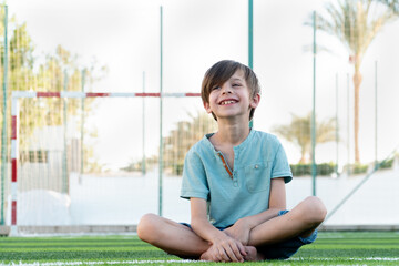 Positive smiling boy sitting on lawn with crossed barefoot legs and looking away 