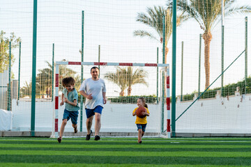 Full body positive man with children running together on sports field to camera during playing football 