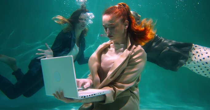 Workplace conflicts. Attractive young red haired businesswoman using laptop underwater, colleagues shouting slow motion.
